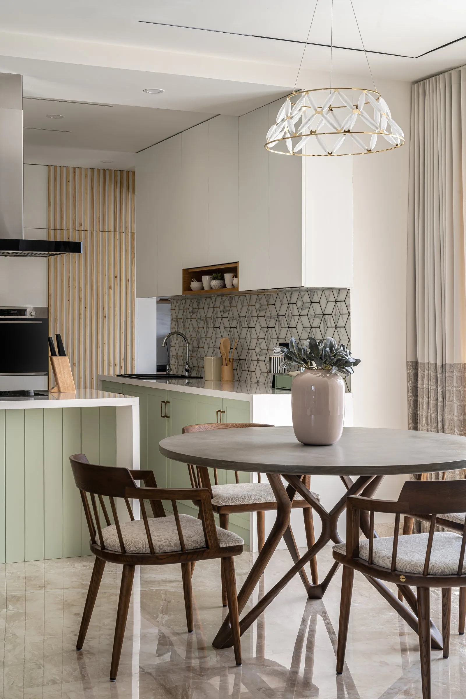 Round walnut dining table under a white geometric pendant, with sage kitchen cabinets behind